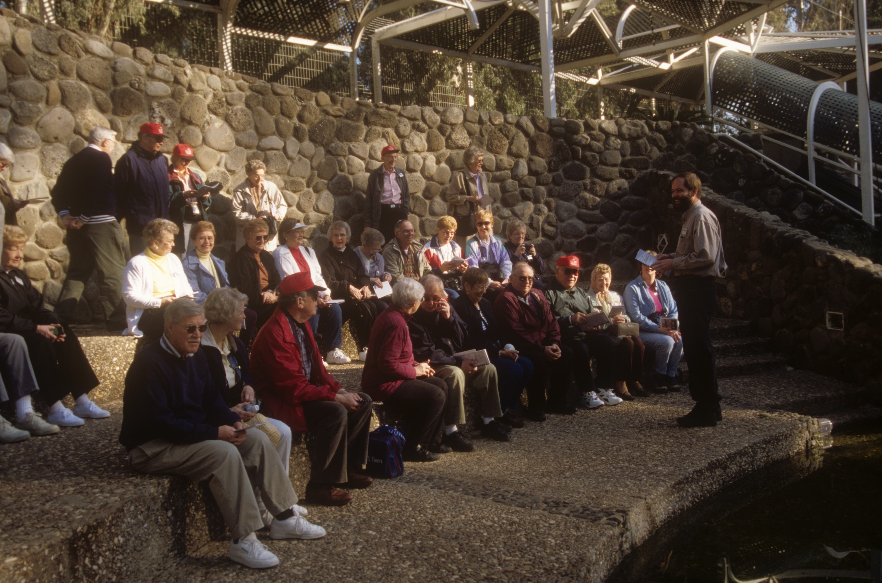 Large group of pilgrims seated outdoors listening to a guide at a historic site in the Holy Land