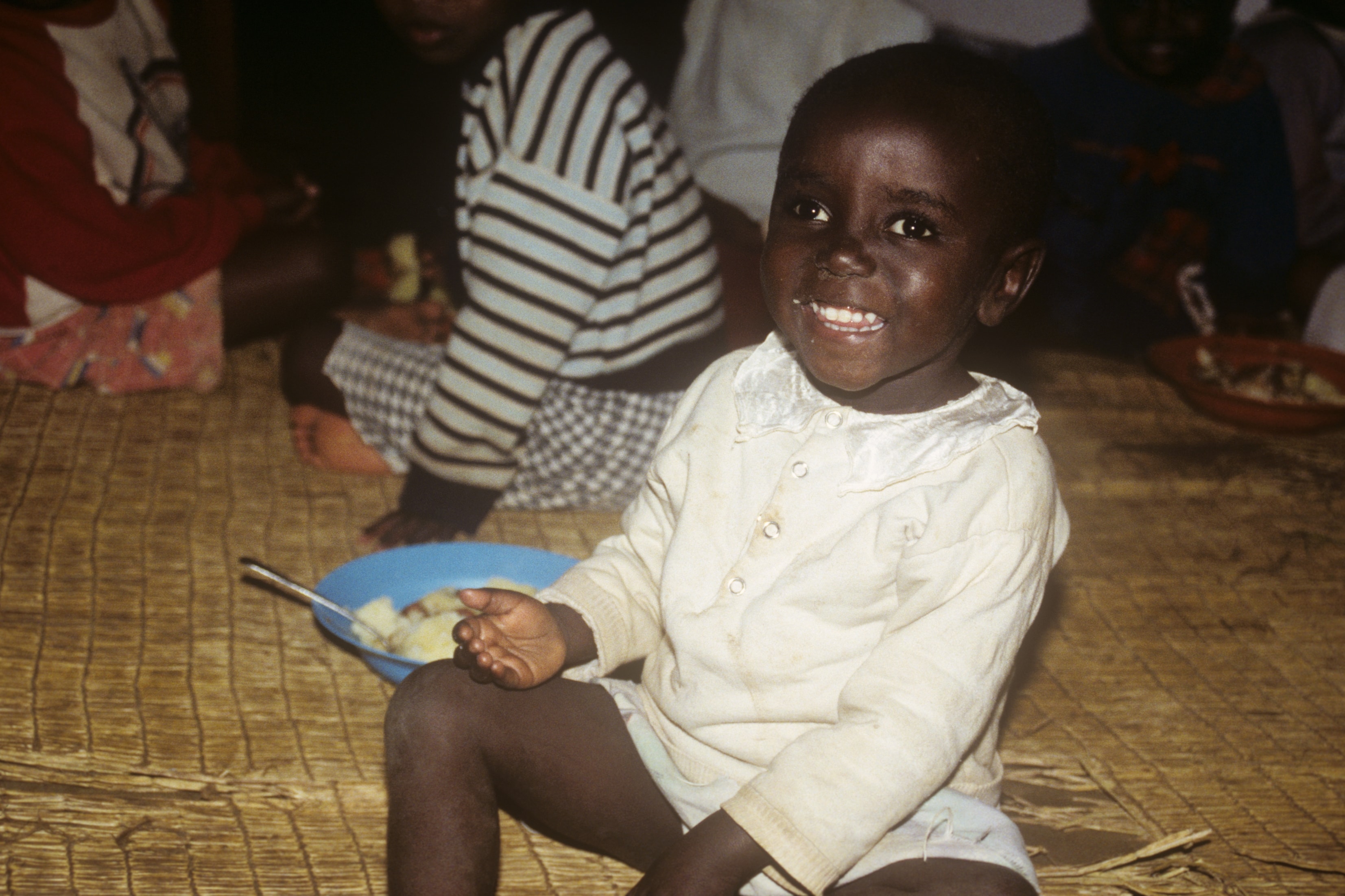 Young child smiling while eating from a bowl on a woven mat in Rwanda