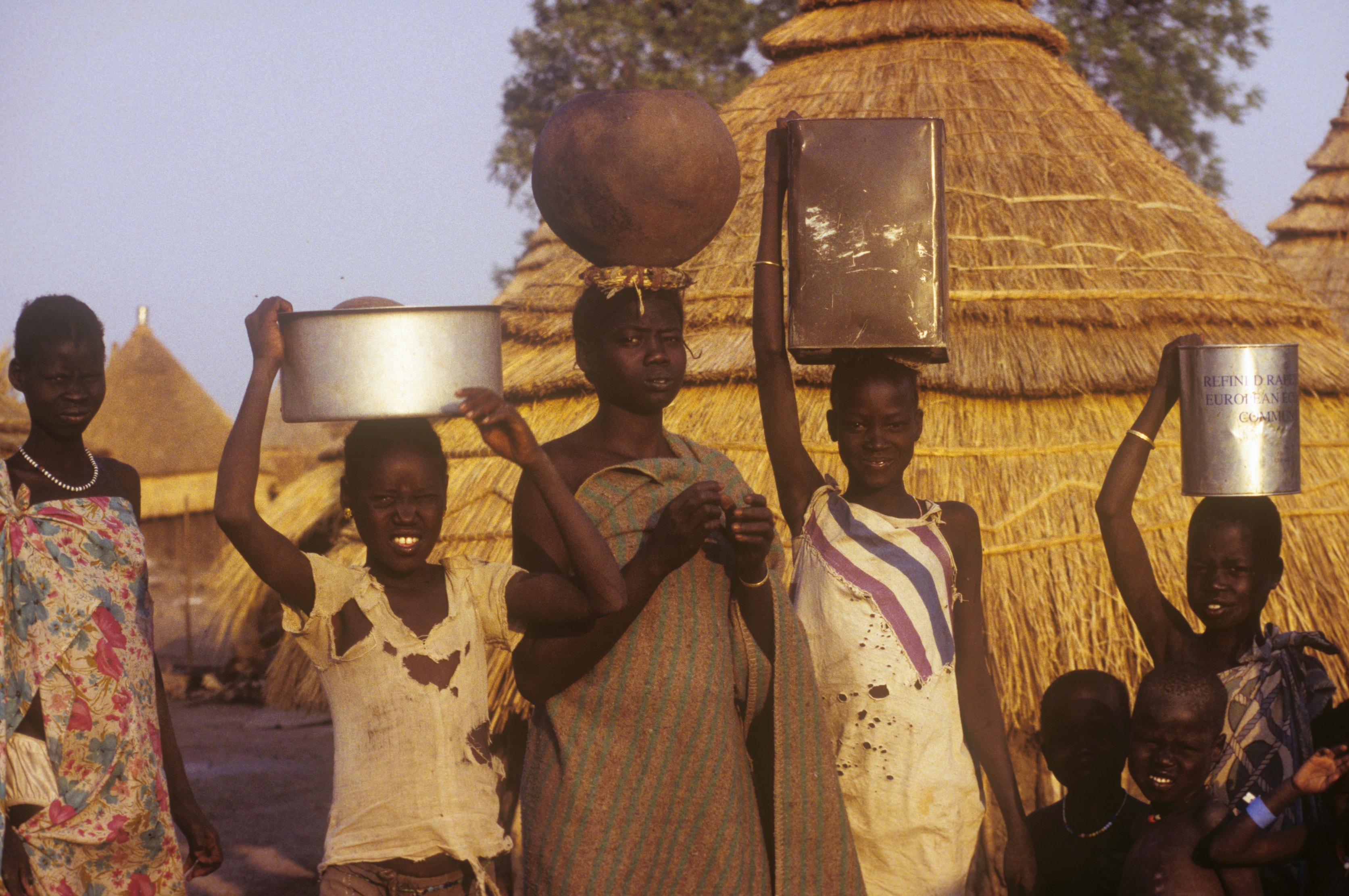 Women and children carrying pots and containers on their heads in front of thatched huts in Sudan